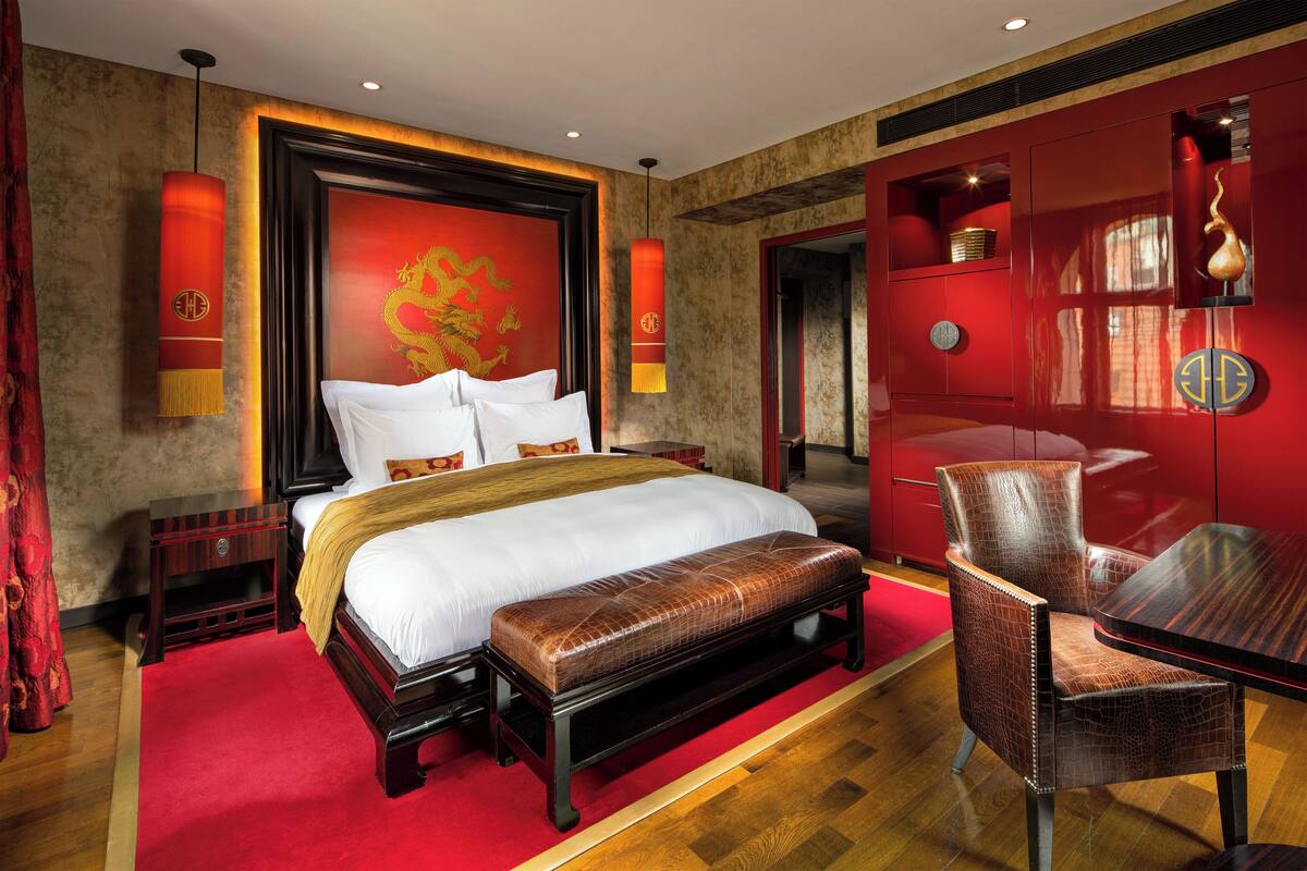 A spacious bedroom with a large bed, a wooden bench, a red accent wall, and a traditional Chinese dragon mural.