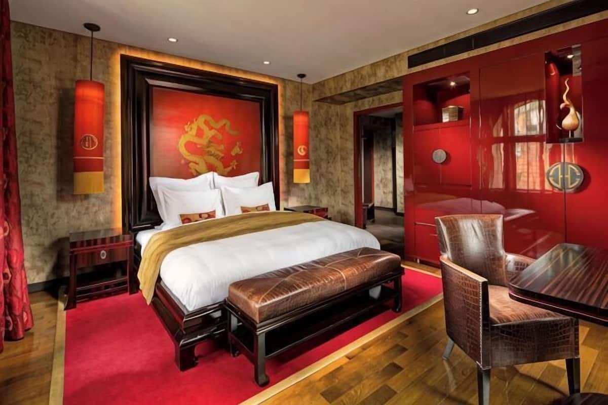 A hotel room with a large bed, a wooden bench, a red wall with traditional decor, and a small table.