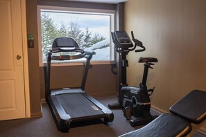 Una habitación con cinta de correr y máquina elíptica, una ventana con vista a árboles y nieve, y una puerta.