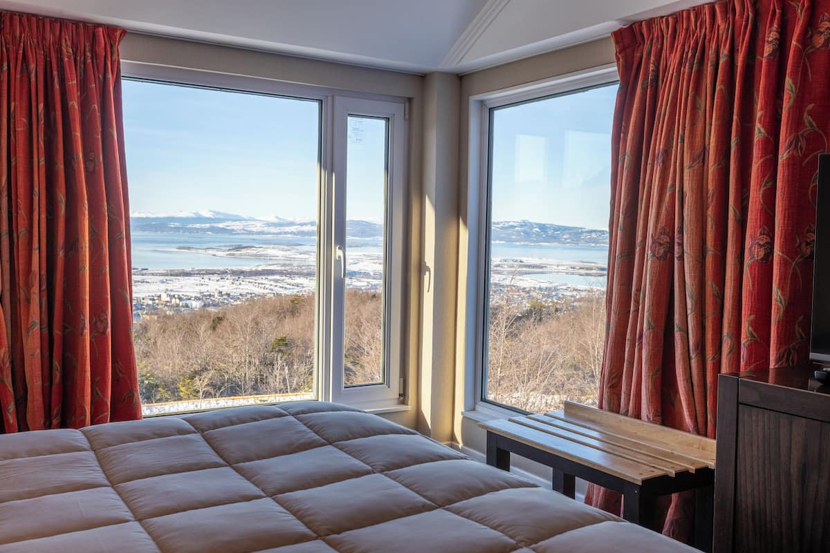 A bedroom with a large bed, a wooden nightstand, and a view of a snowy landscape through two windows.