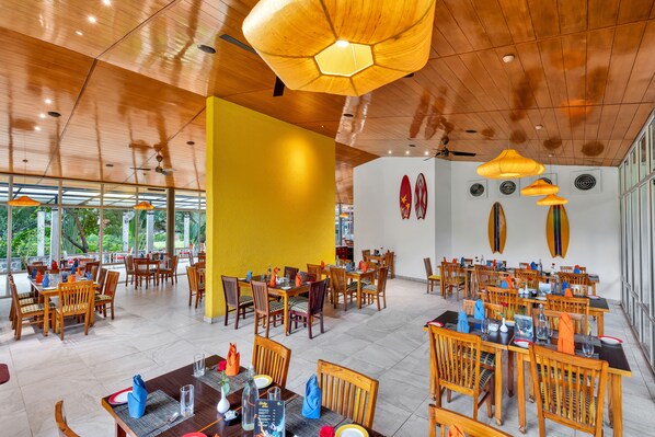 A spacious dining area with wooden tables and chairs, large windows, and a yellow accent wall.