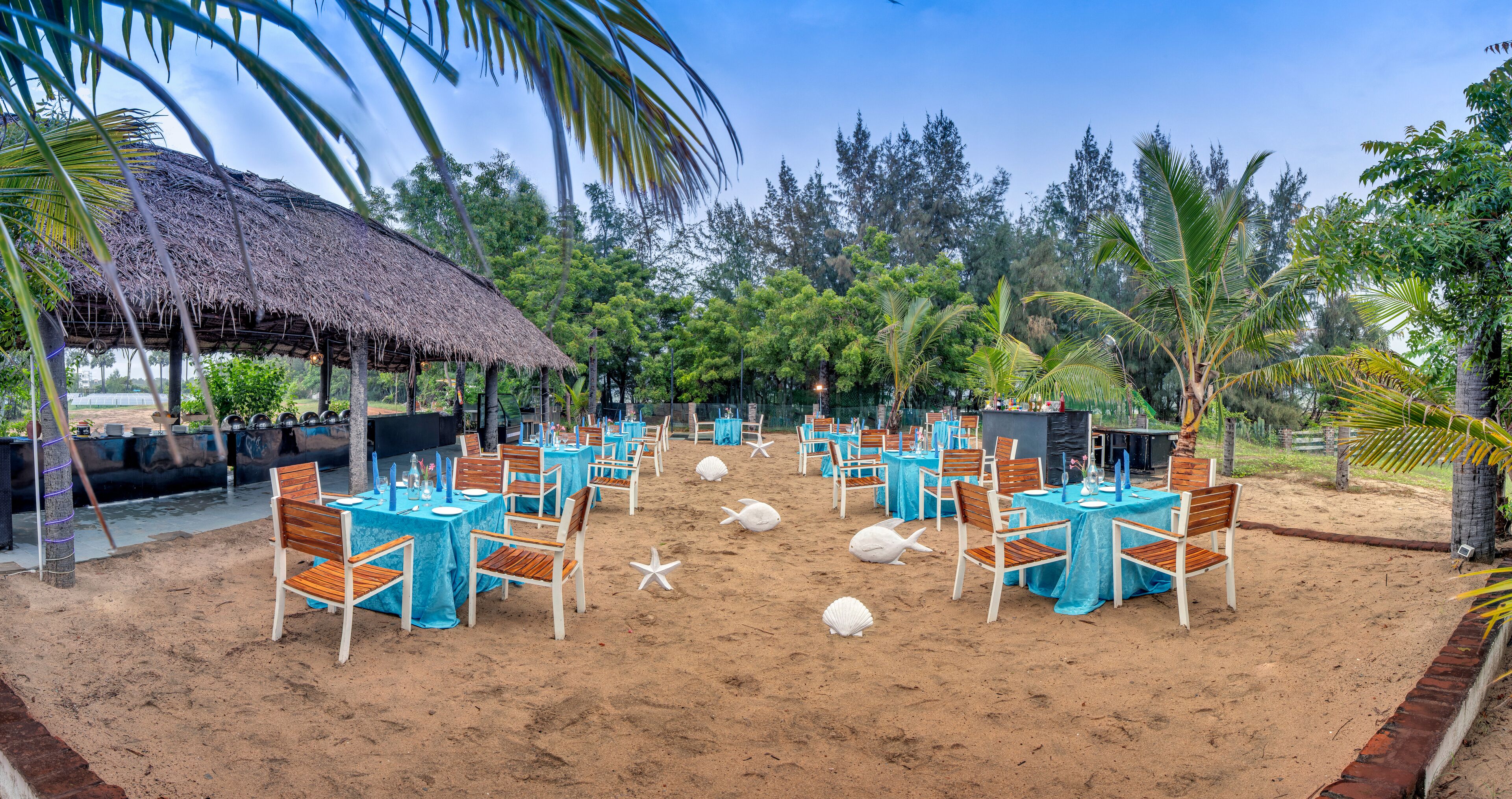 Un área de comedor al aire libre con mesas y sillas, una estructura de techo de paja y un entorno de playa arenosa.