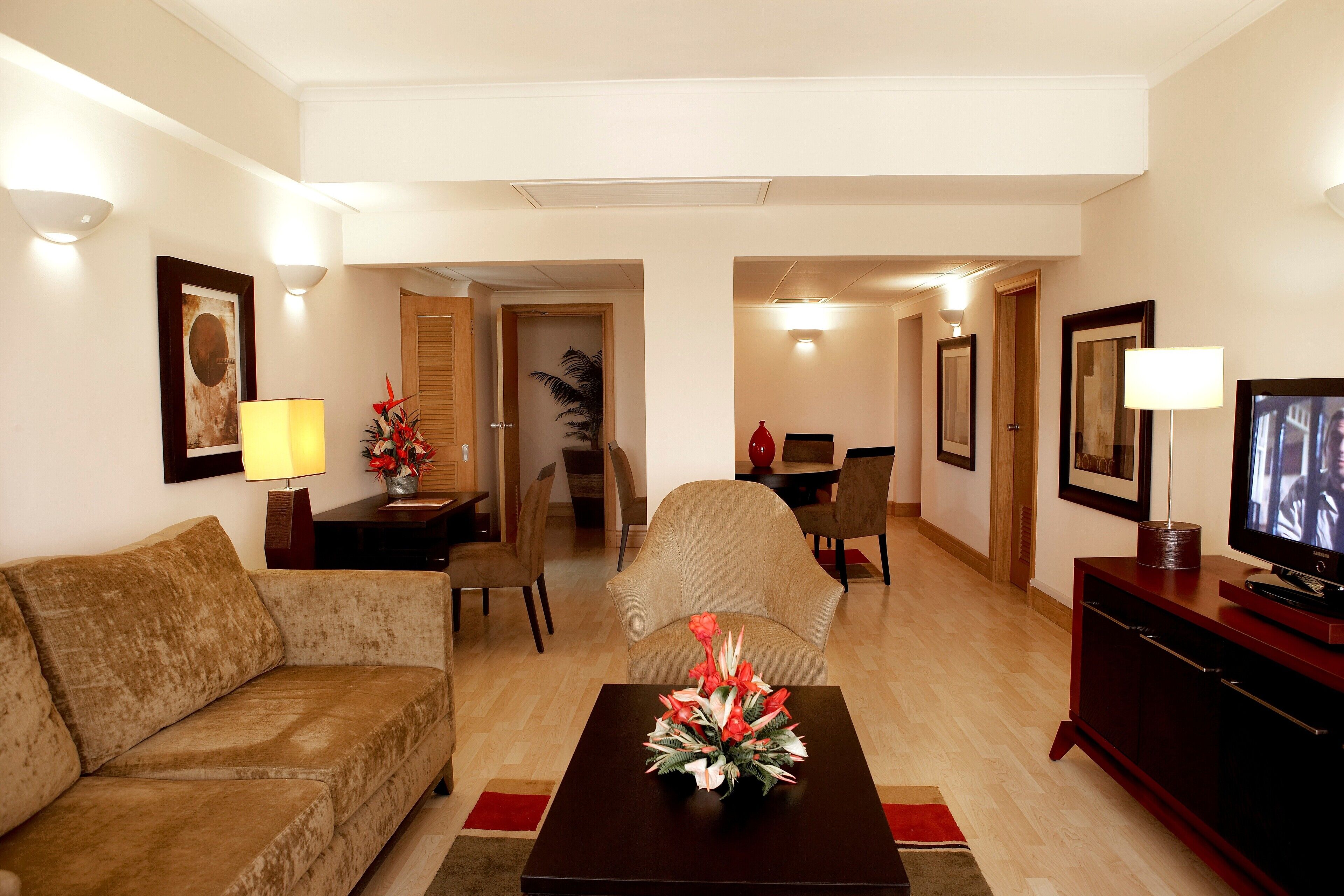 A living room with a sofa, a coffee table with a floral arrangement, a television on a cabinet, and a dining area in the background.