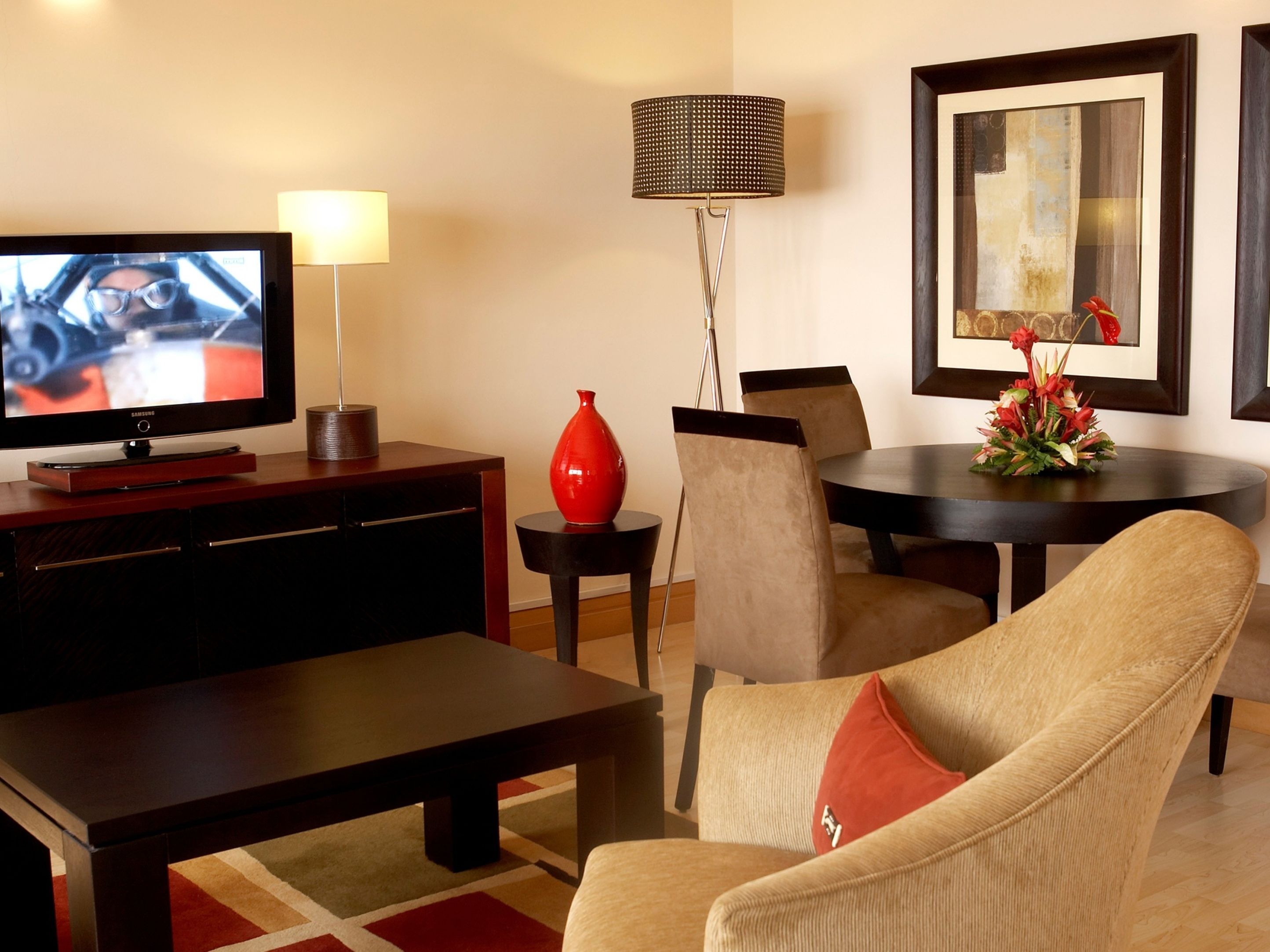 A modern living room with a flat-screen TV, a red vase, a lamp, a dining table, and a sofa with a red pillow.