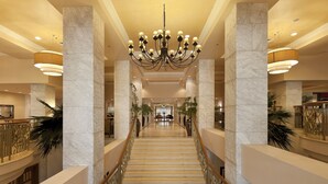 A grand hotel lobby with marble columns, a central chandelier, and a staircase leading to upper floors.