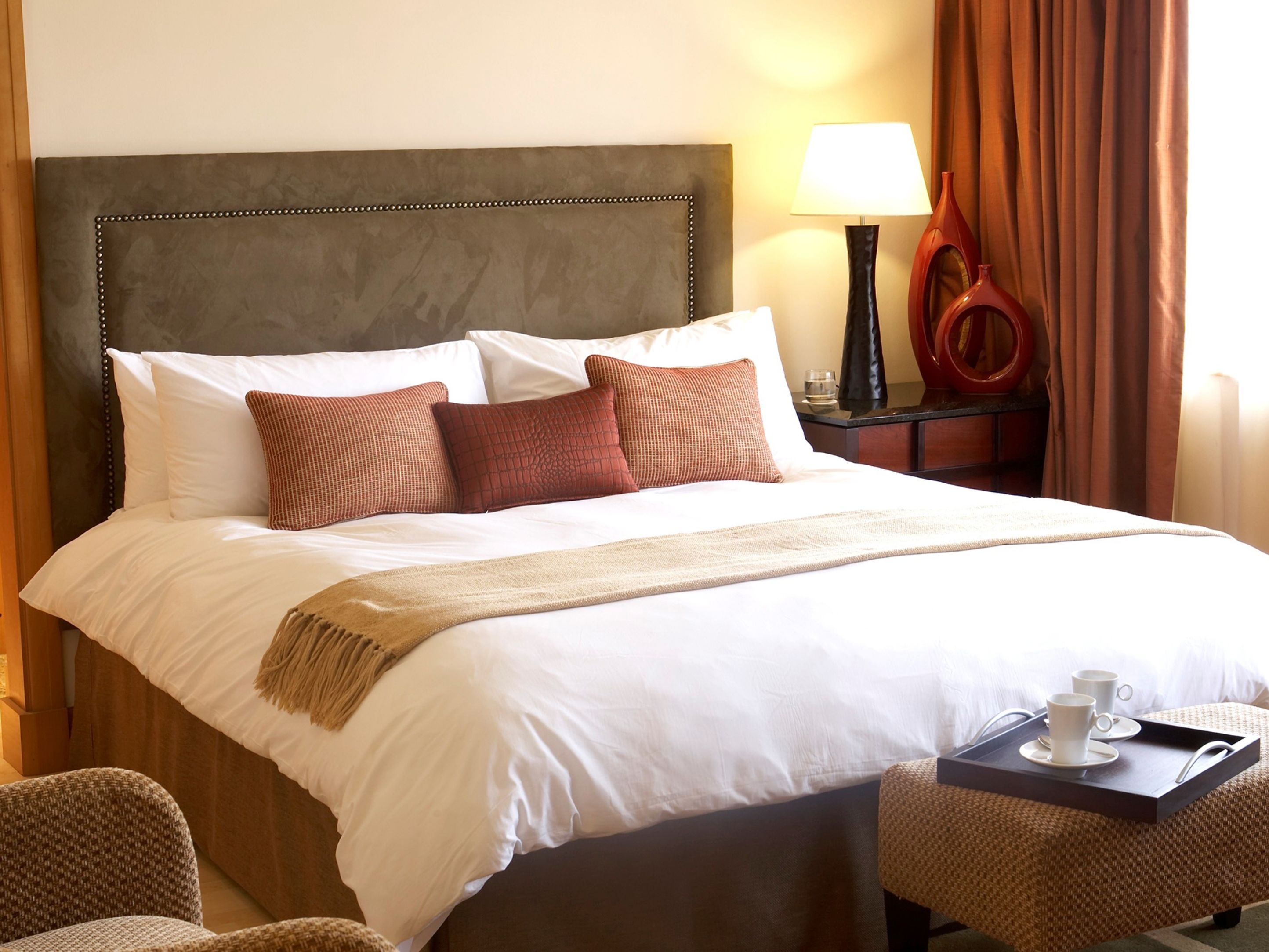 A neatly made bed with white and brown bedding, a bedside table with a lamp, and a tray with cups.