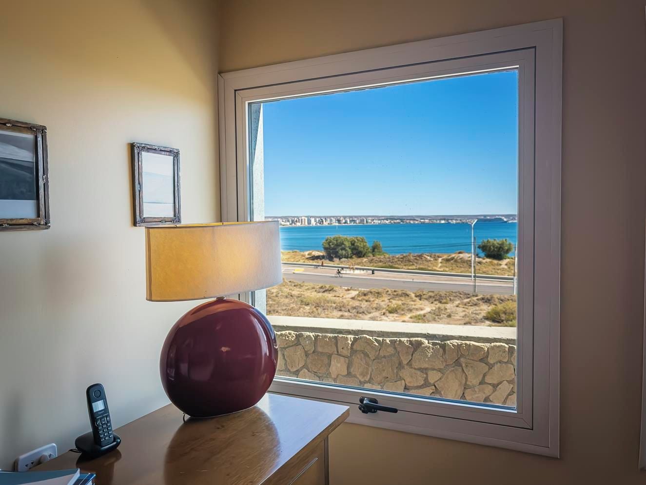 A room with a large window overlooking a coastal view, a lamp, and a telephone on a wooden table.