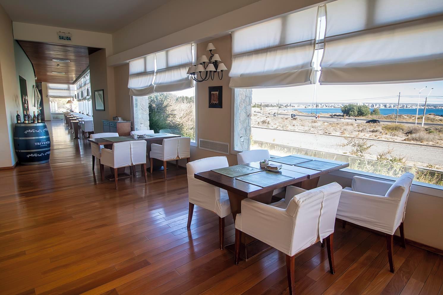 A dining area with wooden flooring, white chairs, and a table set for two. Large windows with blinds offer a view of the beach and road.