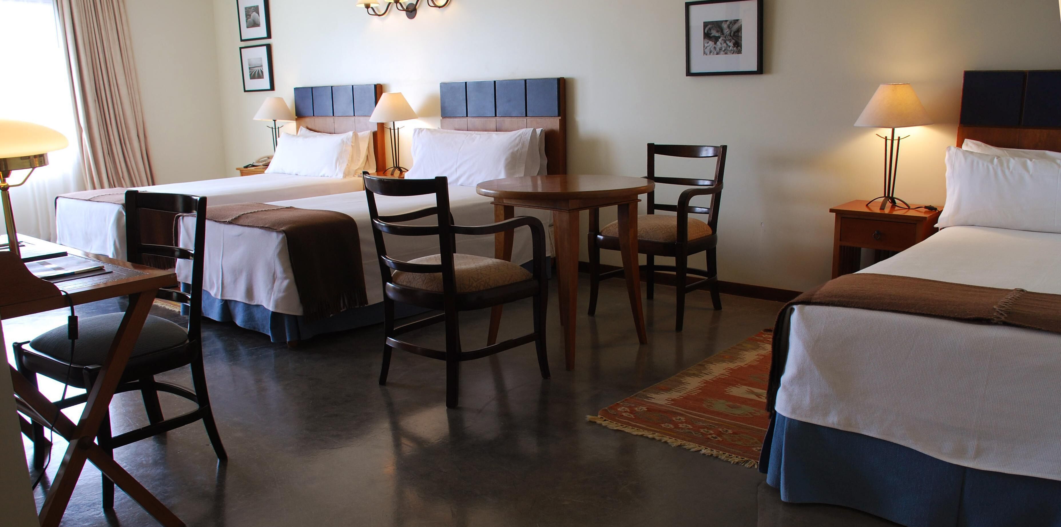 A hotel room with two beds, a desk, a chair, a small table, and a nightstand.