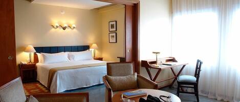 A hotel room with a bed, two chairs, a small table, and a desk. There are wall-mounted lights and a window with curtains.