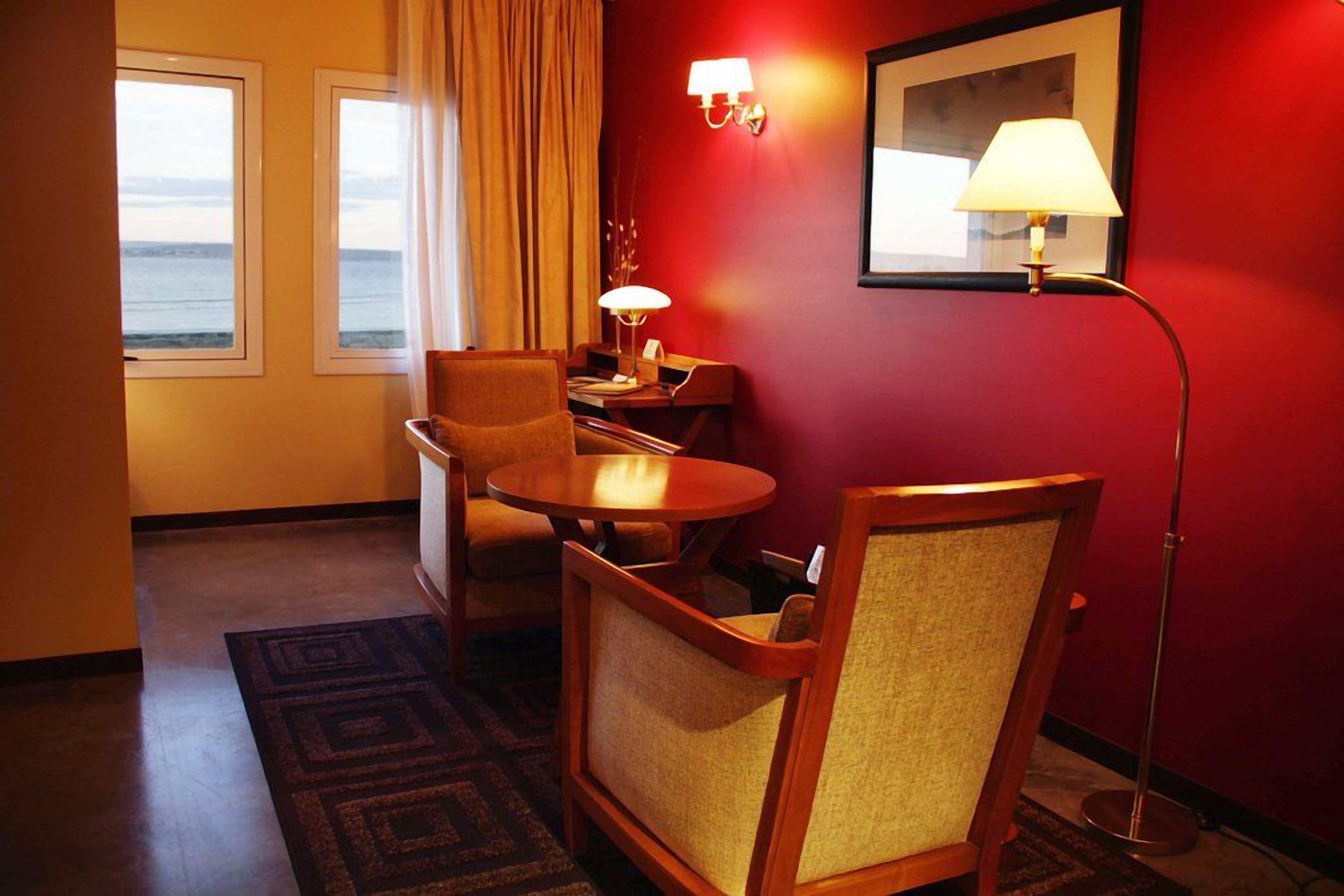 A hotel room with a view of the ocean, a desk with a lamp, two chairs, and a red wall.