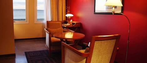 A hotel room with a view of the ocean, a desk with a lamp, two chairs, and a red wall.