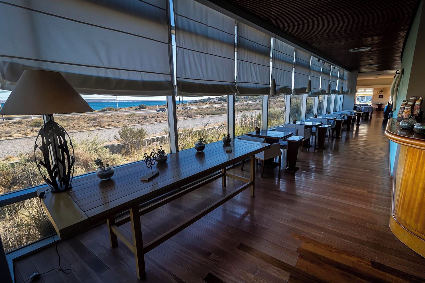 A long dining area with tables and chairs, large windows with blinds, and a view of the landscape.