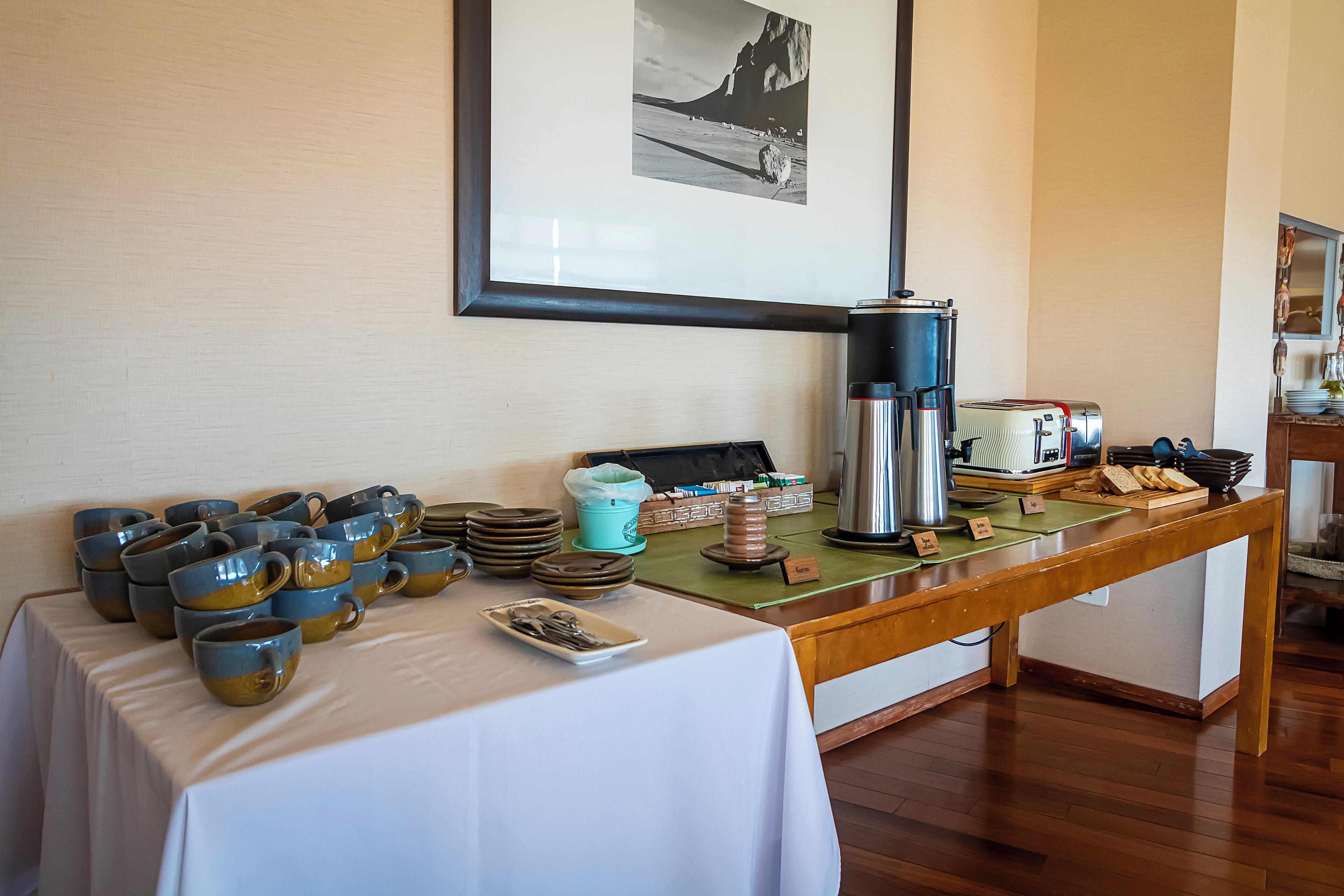 A breakfast buffet with ceramic mugs, a coffee pot, and a toaster on a wooden table.