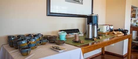 A breakfast buffet with ceramic mugs, a coffee pot, and a toaster on a wooden table.