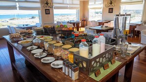 Montaje de bufé con diversos alimentos y salsas, un dispensador de leche y vistas a la playa a través de amplias ventanas.
