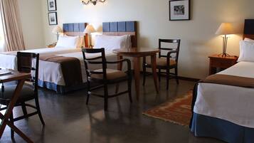 A hotel room with two beds, a desk, two chairs, a small table, and a nightstand with a lamp.