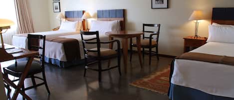 A hotel room with two beds, a desk, two chairs, a small table, and a nightstand with a lamp.