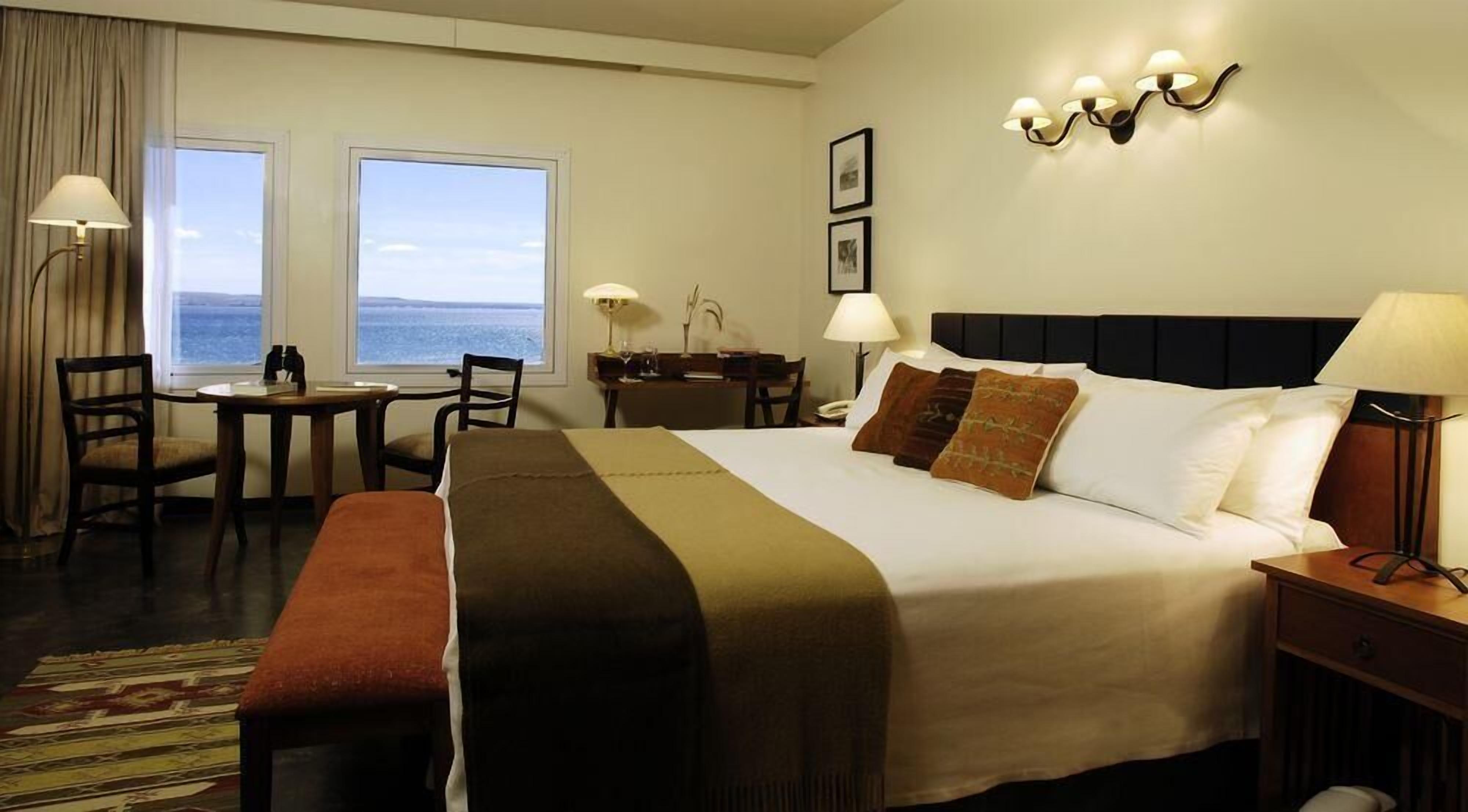 A hotel room with a bed, two chairs, a desk, a lamp, a bench, and a view of the sea.