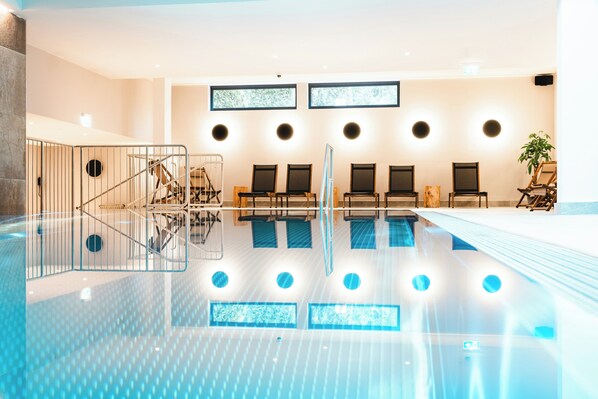 A modern indoor swimming pool with clear water, a seating area with chairs, and a staircase leading to a lower level.