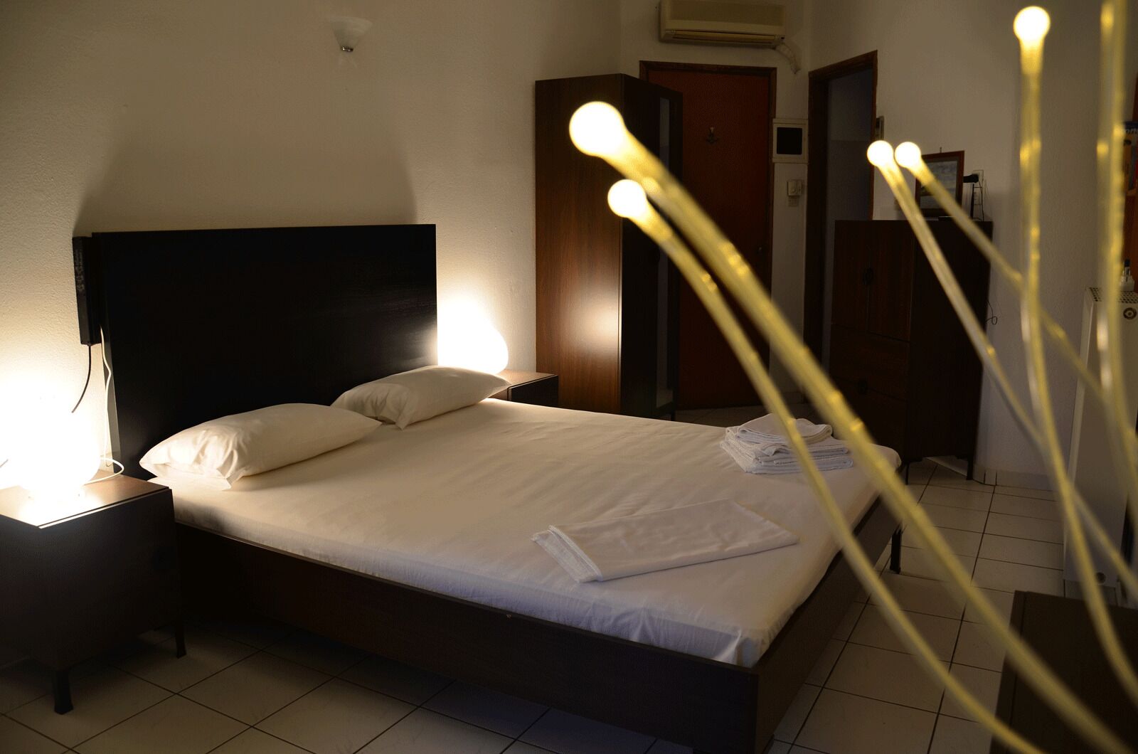 Un lit bien fait, avec des draps blancs, une lampe de chevet et une serviette pliée posée sur le lit.