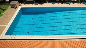 A rectangular swimming pool with clear blue water, surrounded by a tiled deck and a covered area with lounge chairs. - Saron Hotel (Lavreotiki)