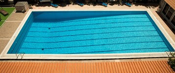 Una piscina rectangular con agua azul cristalina, rodeada por un solárium con baldosas y una zona cubierta con sillones de descanso.