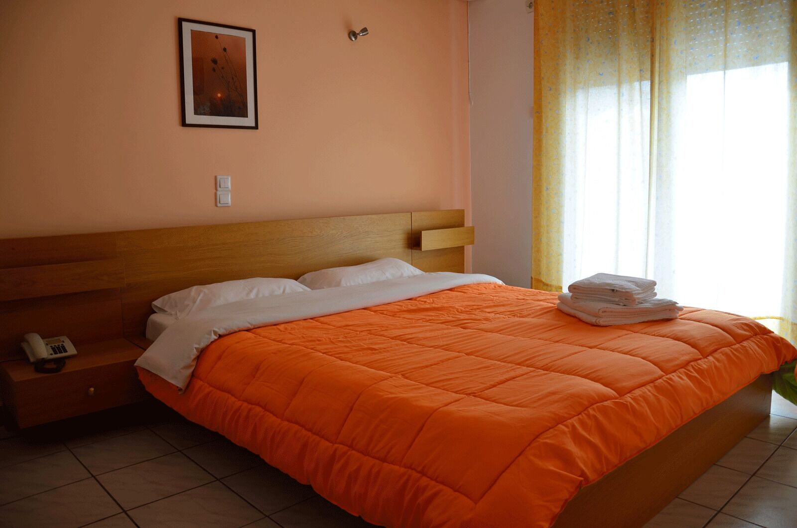 A neatly made bed with an orange blanket, a wooden headboard, a bedside table with a telephone, and a framed picture on the wall.