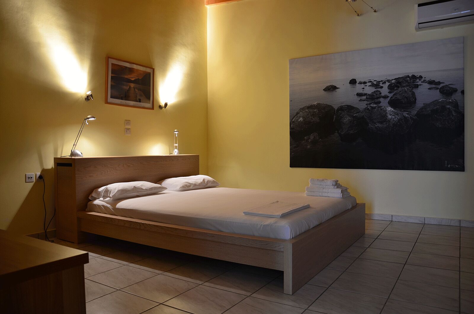 A single bed with a wooden headboard, two bedside lamps, a framed picture on the wall, and a large abstract painting.