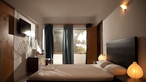A modern bedroom with a large bed, a flat-screen TV, a bedside table with a lamp, and a view through a sliding door to the outside. - Saron Hotel (Lavreotiki)