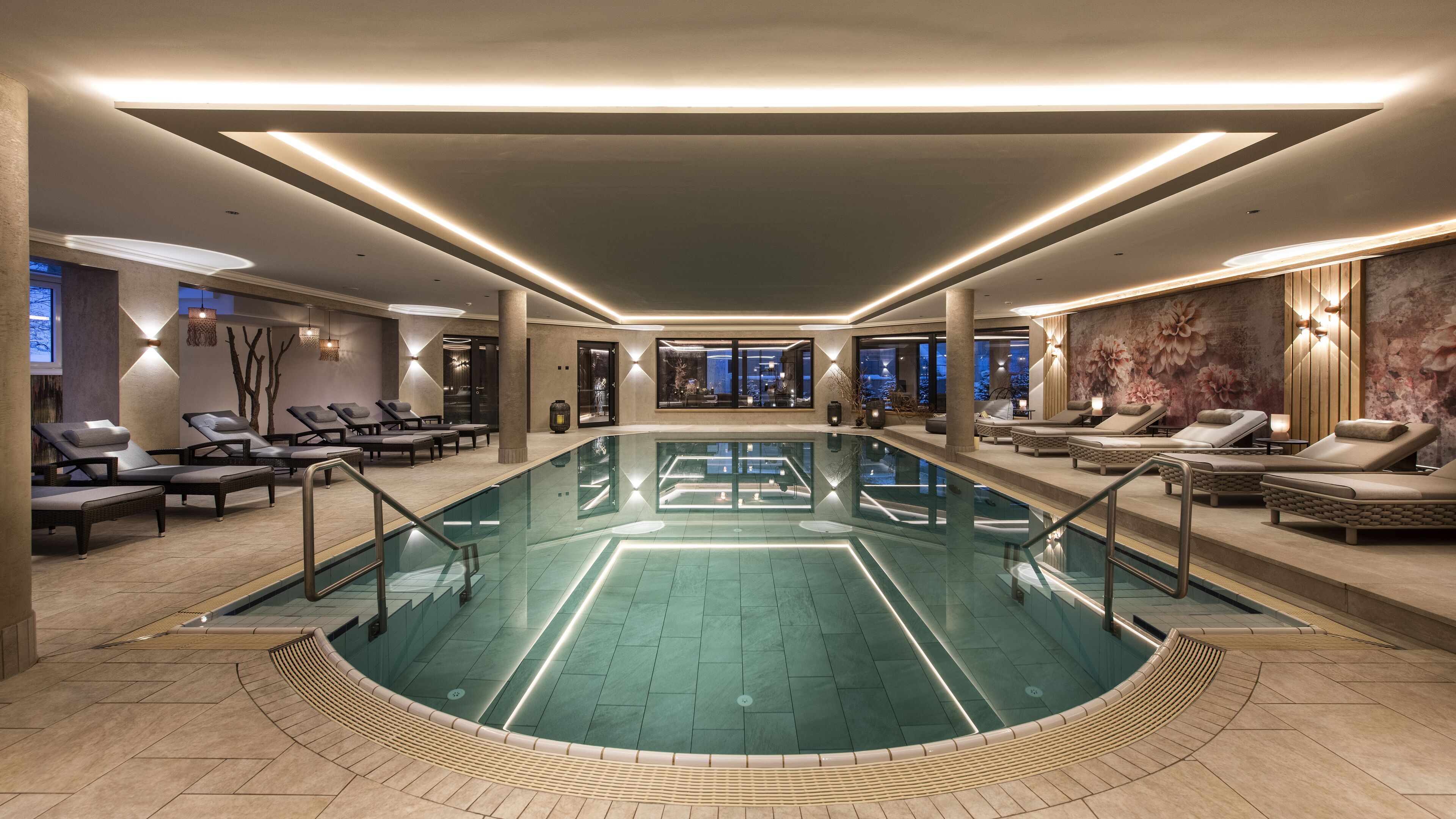 A large indoor swimming pool with lounge areas on both sides, a view of the cityscape through large windows, and modern lighting fixtures.
