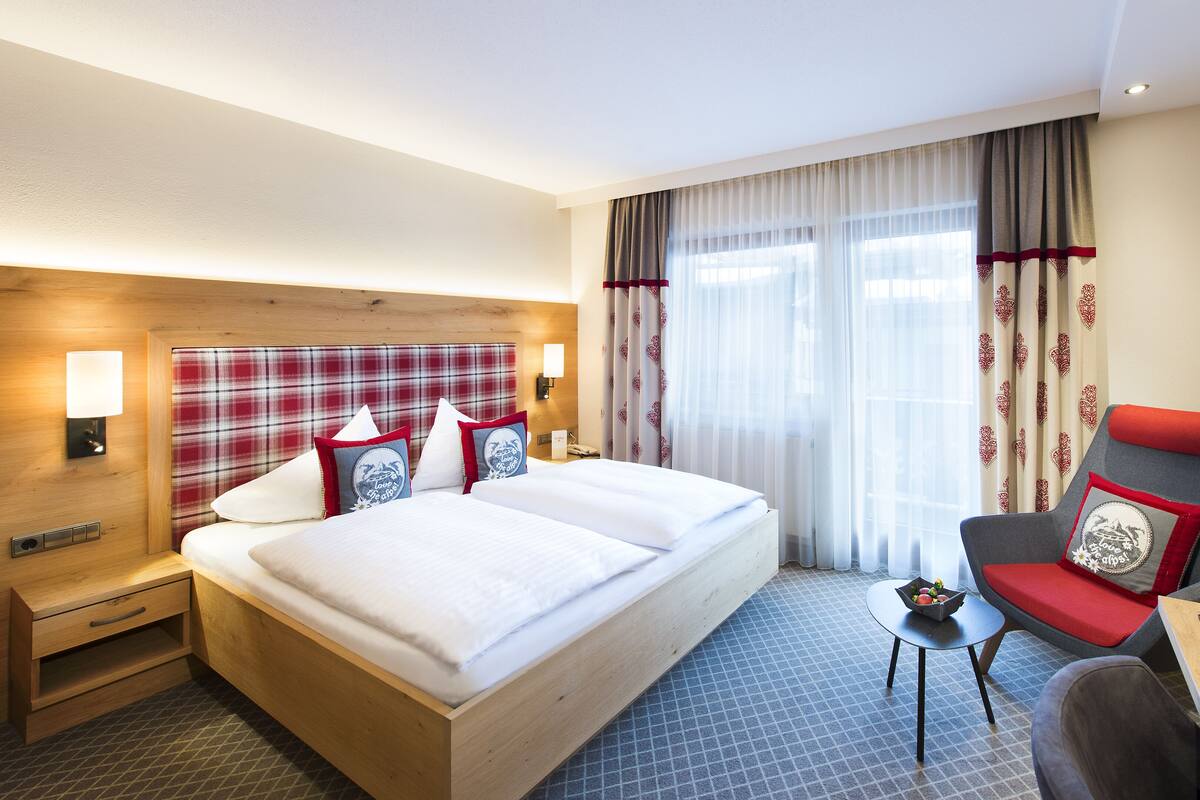 Hotellihuone, jossa on puinen sängynpääty, valkoisilla lakanoiden peitetty sänky, punavalkoinen ruutukuviollinen sängynpääty, pieni pöytä, jossa on kulho täynnä hedelmiä, ja punaisilla tyynyillä varustettu tuoli.