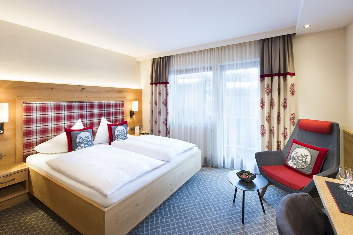 Habitación de hotel con una cama grande, cabecera de madera, una mesita con un frutero y una silla con cojín rojo.
