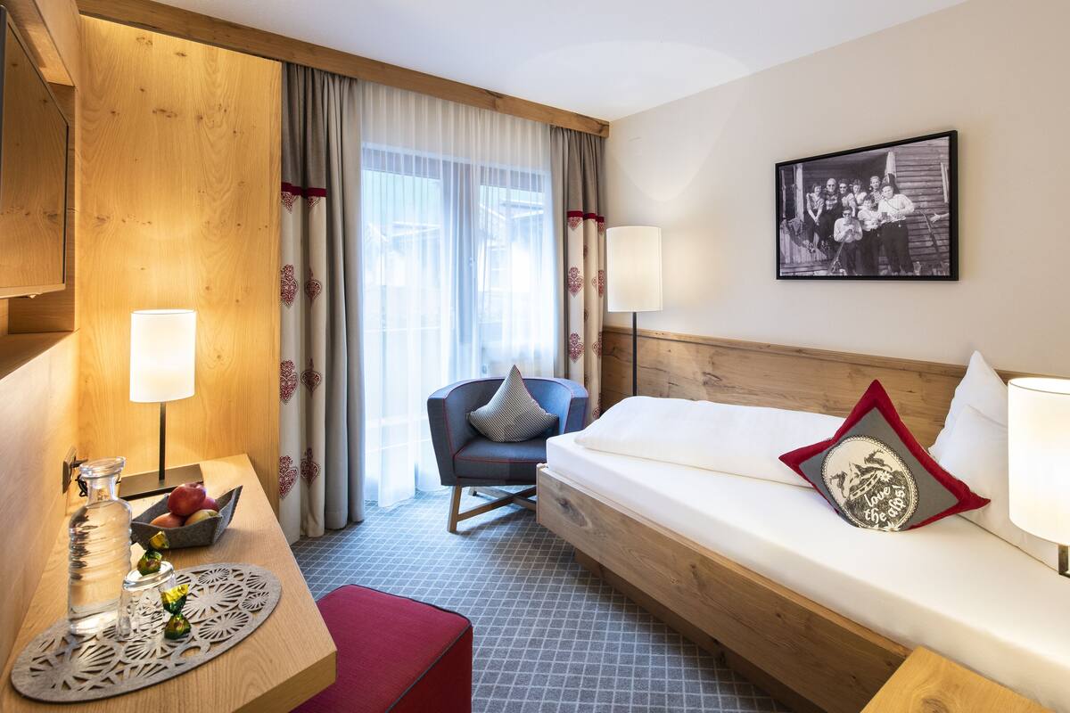 Hotellihuone, jossa on puinen sängynpääty, valkoisilla lakanoiden peitetty sänky, lamppu varustettu työpöytä, pehmusteella varustettu tuoli ja seinällä oleva kehystetty valokuva.