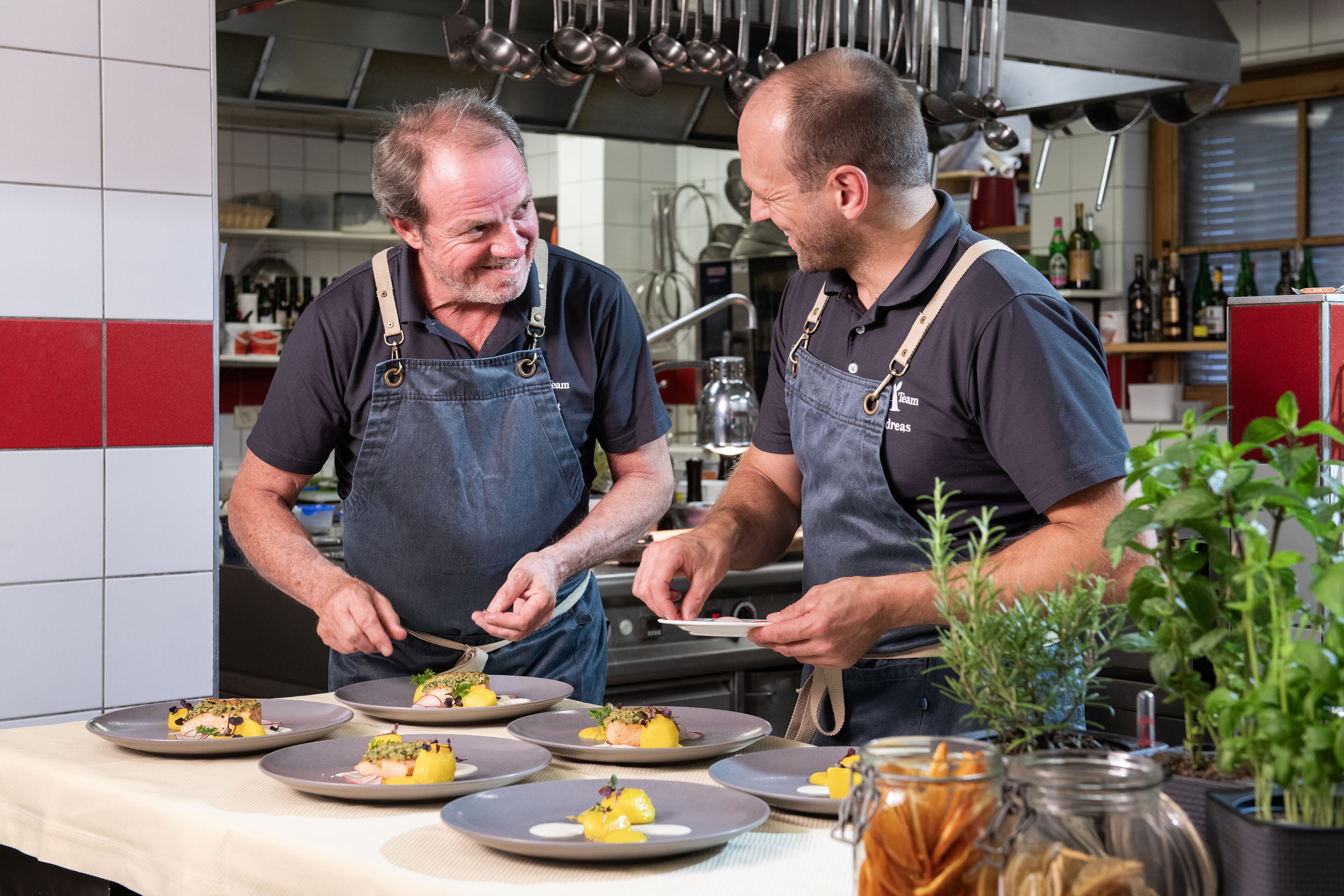 Two chefs preparing dishes in a kitchen with tiled walls and various cooking utensils.