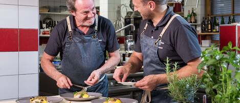Dois chefs preparando pratos em uma cozinha com paredes azulejadas e diversos utensílios de cozinha.