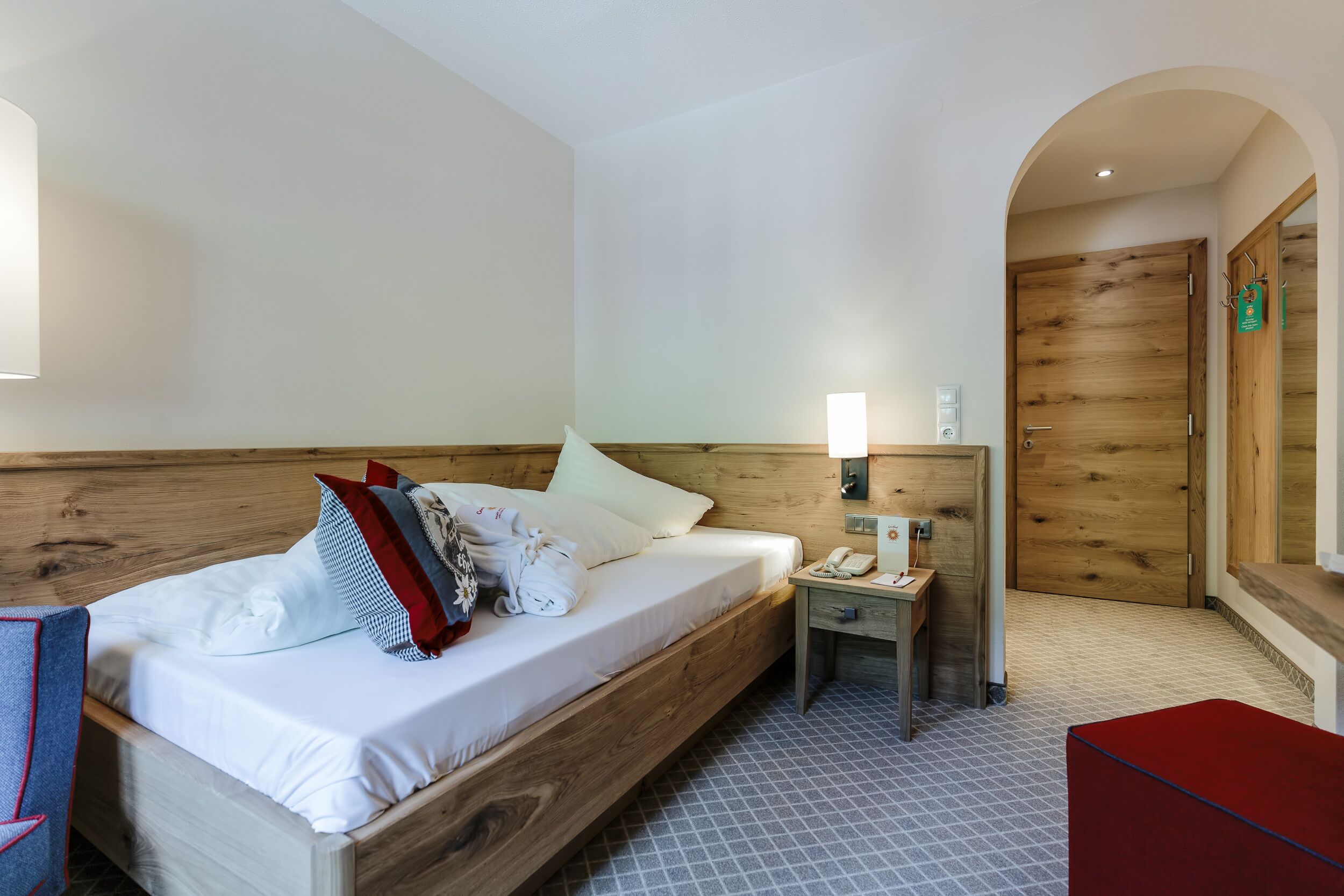 Una habitación de hotel con una cama de madera, una mesita de noche con una lámpara, un teléfono y un banco rojo.