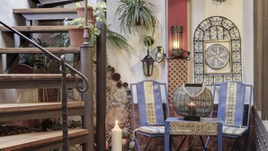 Una escalera con escalones de madera y barandilla de metal, una silla azul con diseños elaborados, una vela blanca grande y una pared decorativa con un arco de metal y un azulejo enmarcado.