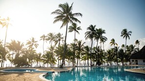 Una piscina rodeada de palmeras, sillas de descanso y un edificio con techo de paja.