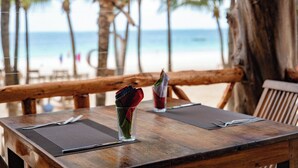 Een houten tafel met een glas met een gestreept drankje, een servet en een menukaart, op een strand met palmbomen en de oceaan op de achtergrond.