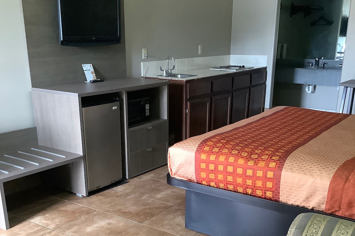 A compact hotel room with a bed, a small kitchenette, a flat-screen TV mounted on the wall, and a bathroom with a mirror and sink.