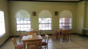 Un espace repas avec des tables et des chaises en bois, de grandes fenêtres équipées de stores blancs et un sol carrelé.