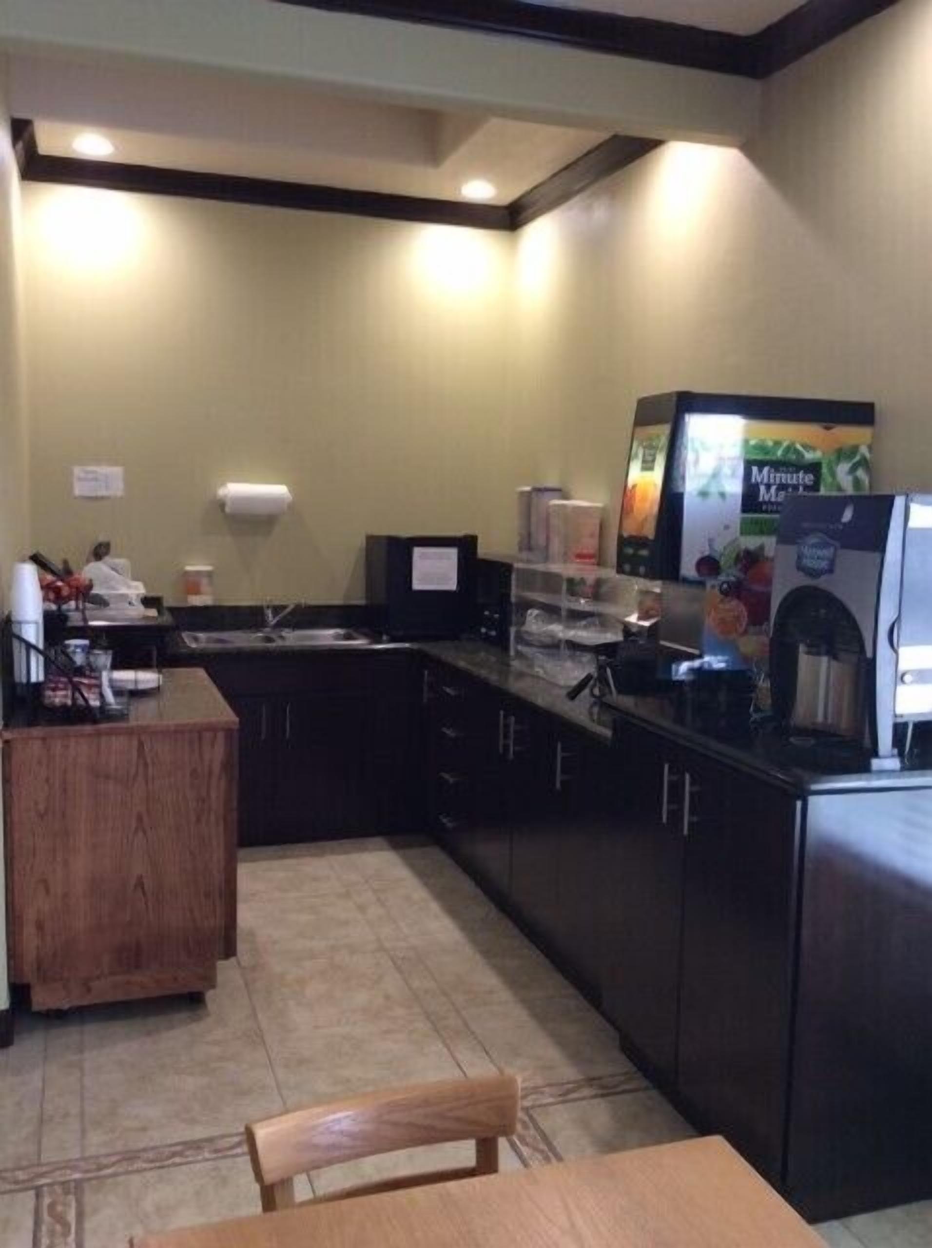 A small kitchenette with a coffee machine, a microwave, and a refrigerator. There is a wooden table and a chair in the foreground.