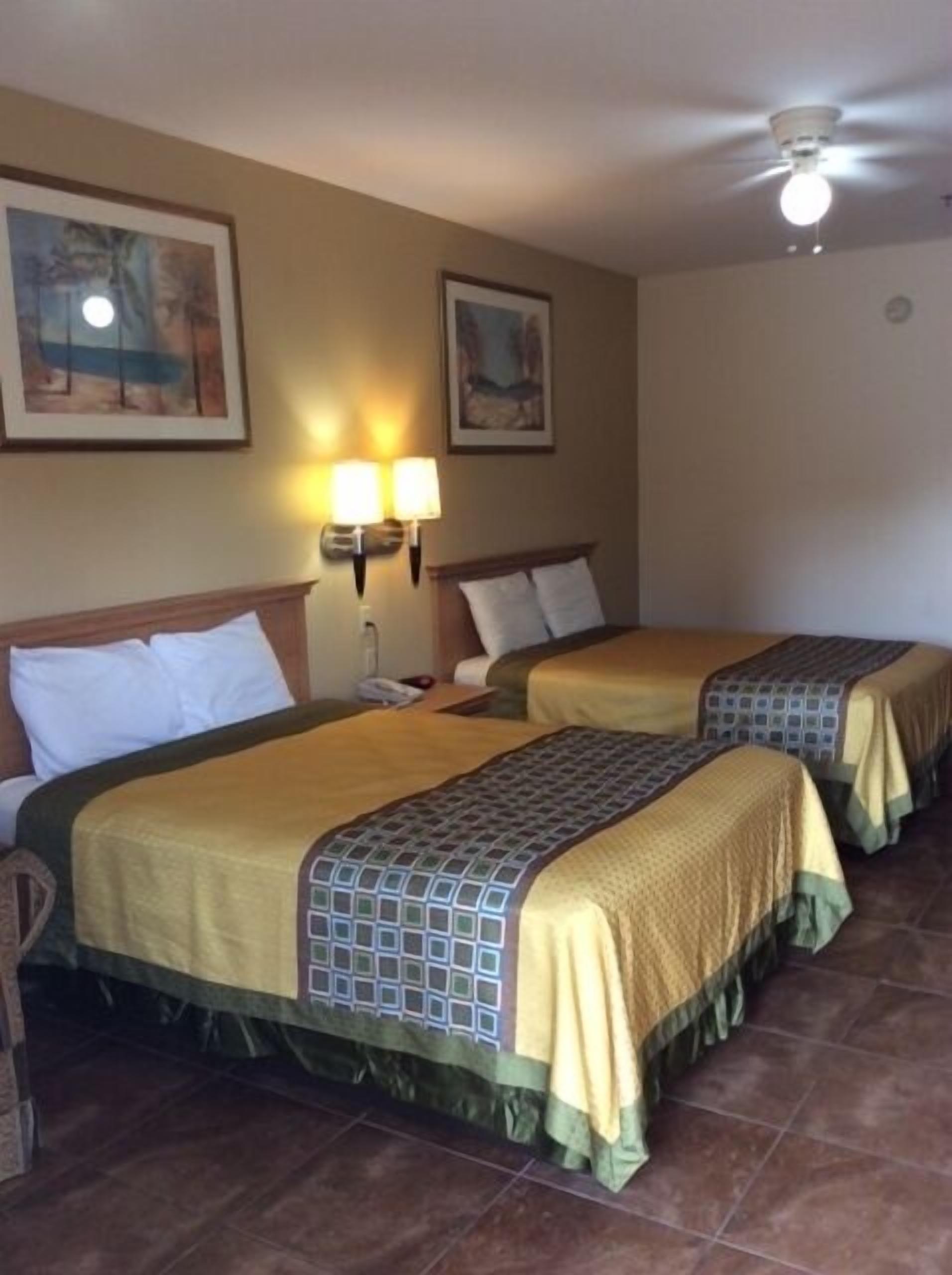 Habitación de hotel con dos camas, ventilador de techo, lámparas fijadas a la pared y cuadros enmarcados.