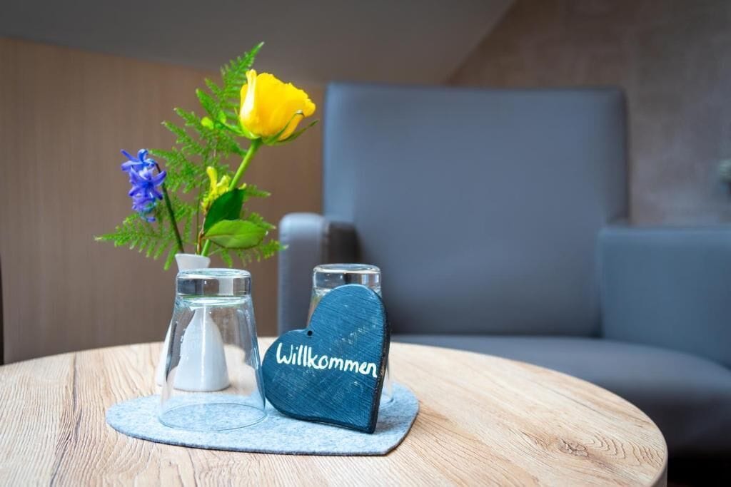 A round wooden table with a glass vase containing a yellow rose and blue flowers, a small glass container, and a heart-shaped coaster with 'Willkommen' written on it.