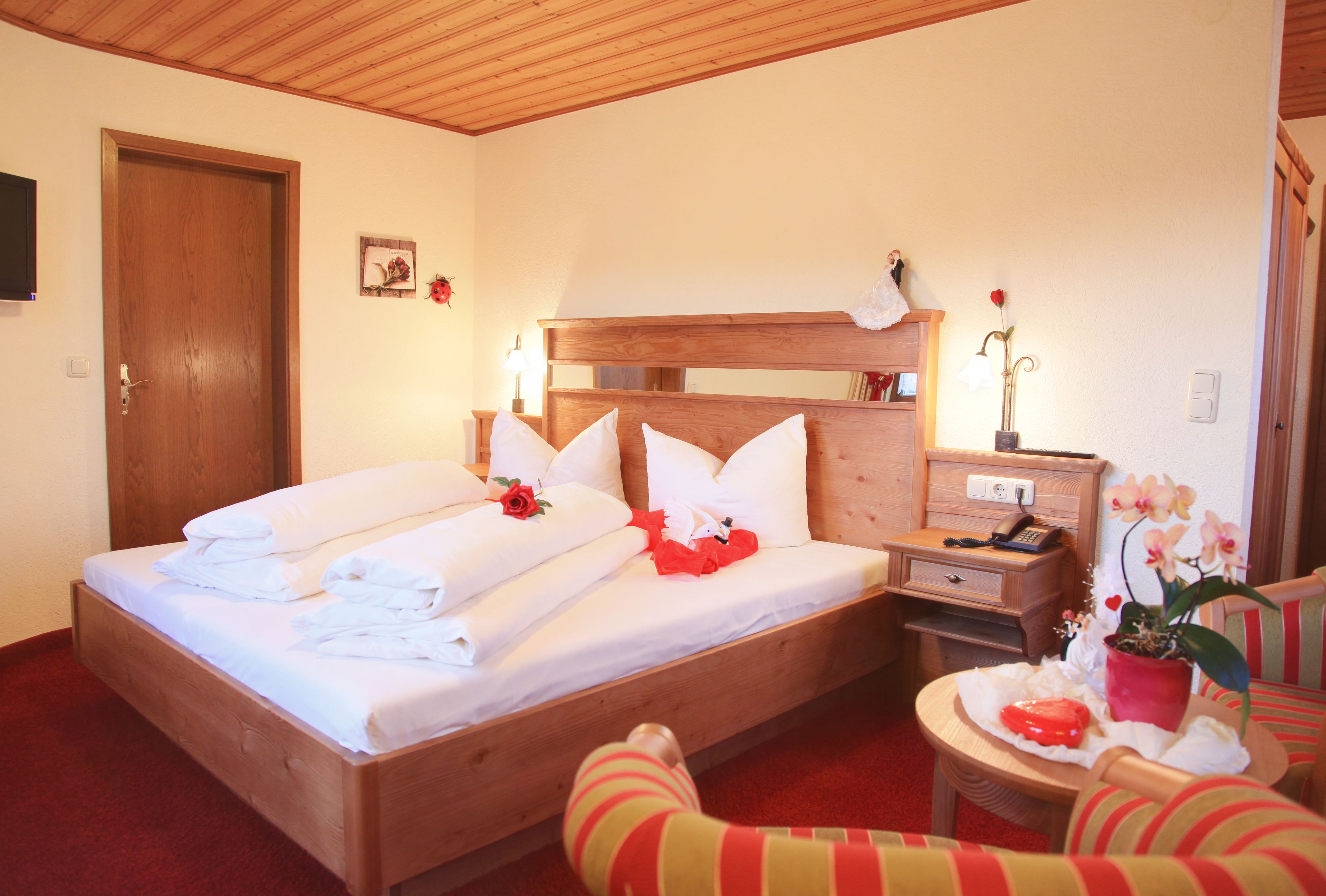 Un letto rifatto con lenzuola e cuscini bianchi, una testiera in legno, un comodino con una lampada, un tavolino con una pianta in vaso e una poltrona a righe.
