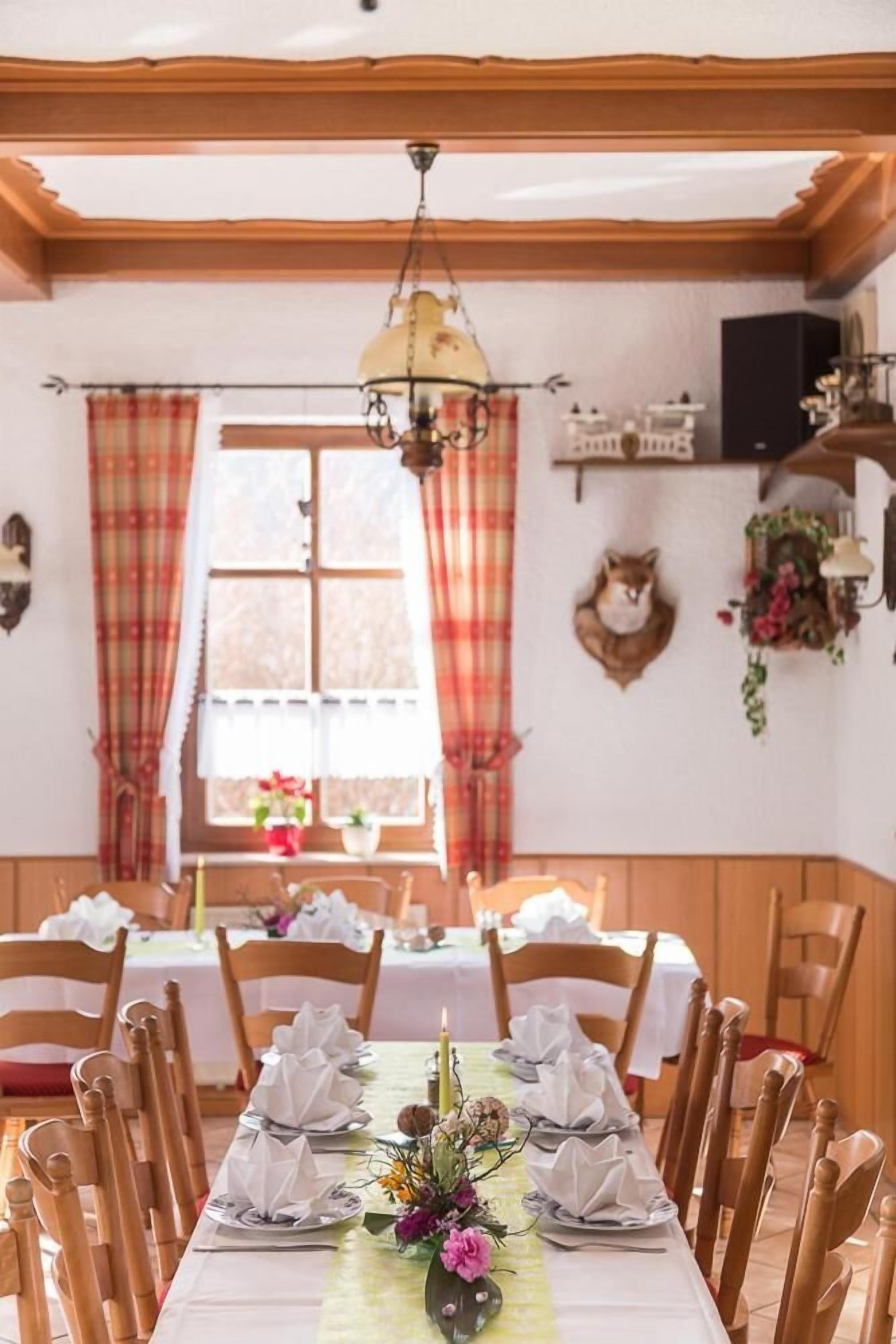 A dining area with a long table set for a meal, wooden chairs, a chandelier, plaid curtains, and a mounted animal head on the wall.