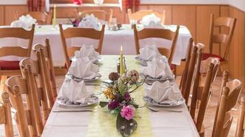 A dining area with a long table set for a meal, wooden chairs, a chandelier, plaid curtains, and a mounted animal head on the wall.