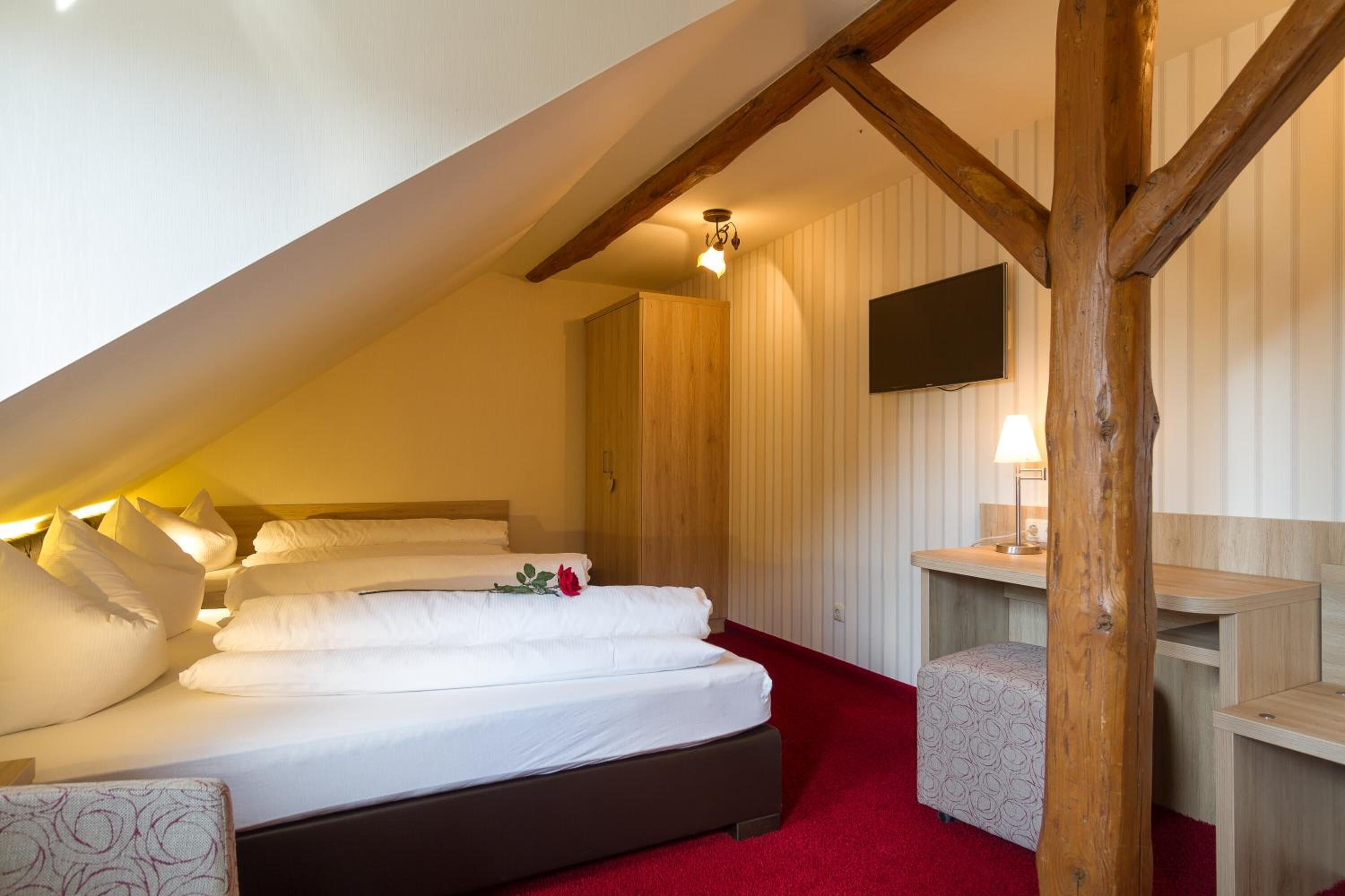 Une chambre avec un plafond en pente, un lit recouvert de linge de maison blanc, une table de chevet en bois et un téléviseur fixé au mur.