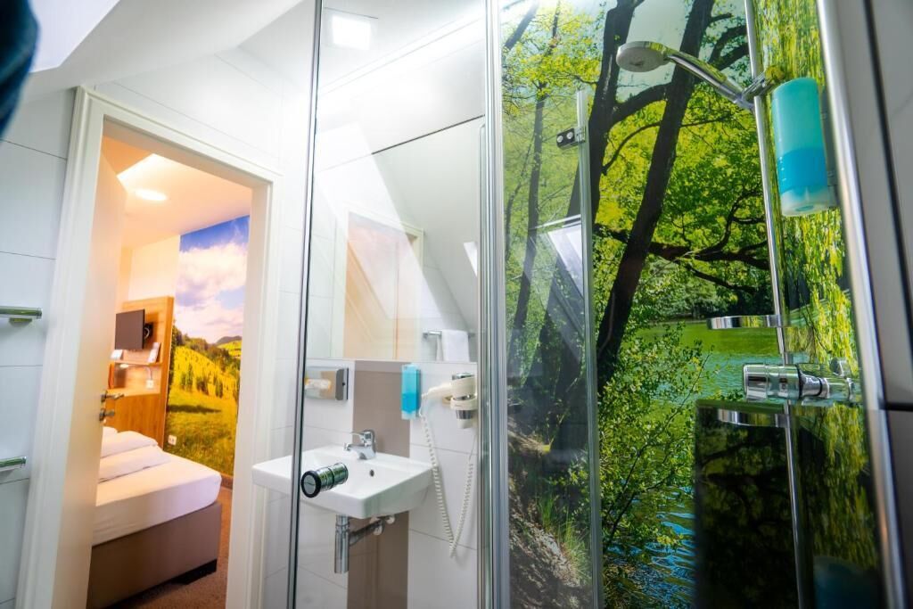 Un baño moderno con cabina de ducha de vidrio, un lavabo y una cama visible a través de una puerta abierta.